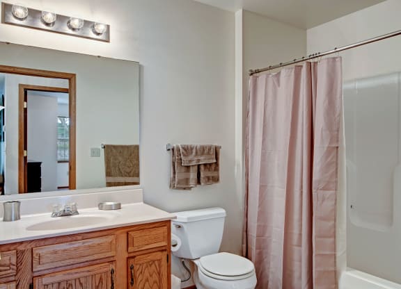 Small bathroom at Arbor Ridge Apartments with wooden vanity, sink, and large mirror. A toilet is beside it, with beige towels hanging above. A pink shower curtain encloses the tub.