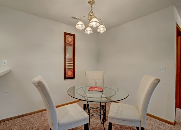 Dining area in Arbor Ridge Apartments featuring a round glass table with three upholstered chairs, a chandelier with five lights above the table, white walls, a decorative mirror on one wall, and light brown carpeted flooring.