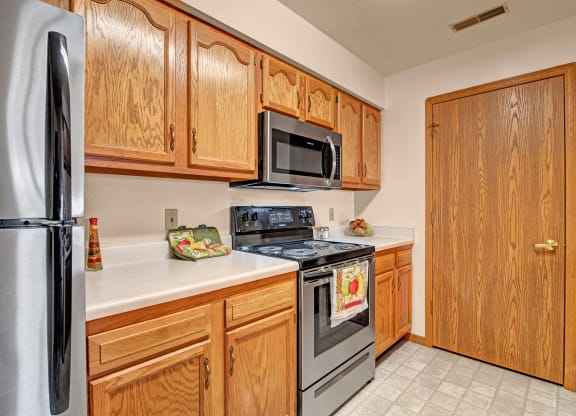 A modern kitchen with wooden cabinets, stainless steel appliances, and white countertops. A decorative towel hangs on the stove, adding a cozy touch. Arbor Ridge Apartments