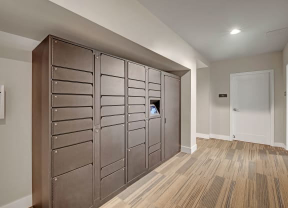 A hallway with a row of dark, numbered parcel lockers and a “Rent Drop” box on the left wall. Neutral tones and a closed white door at River House Apartments.