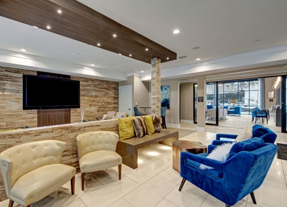 Modern lobby with a sleek, contemporary design. Features cream chairs, a plush blue sofa, colorful cushions, wood accents, and a large TV at River House Apartments.