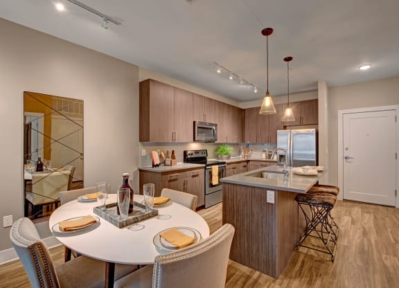 Modern kitchen with sleek wood cabinets, stainless steel appliances, island with barstools, and a round dining table set for four. Warm lighting and neutral tones create a cozy atmosphere at River House Apartments.