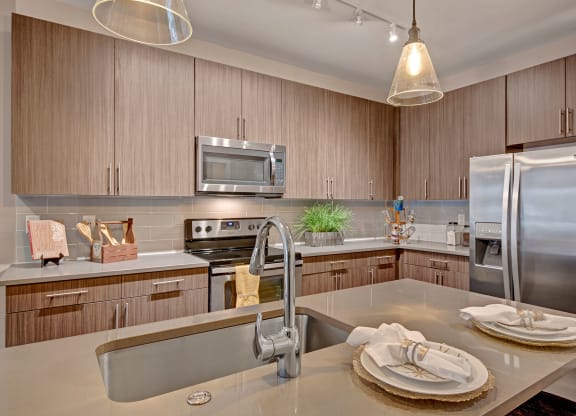 Modern kitchen with wood cabinets, stainless steel appliances, and a double-door fridge. Central island has a sink and two place settings with napkins. Warm lighting creates an inviting atmosphere at River House Apartments.