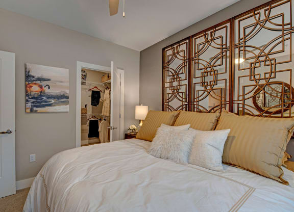 Elegant bedroom with a large, pillowed bed, gold accents, and artistic wall panels. Open closet visible, adding a warm, inviting feel at River House Apartments.