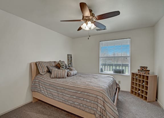 Bedroom with a cozy striped bed under a ceiling fan. A window with blinds lets in light, and a wooden shelf holds patterned boxes, creating a warm, inviting atmosphere at St. James Place Apartments.