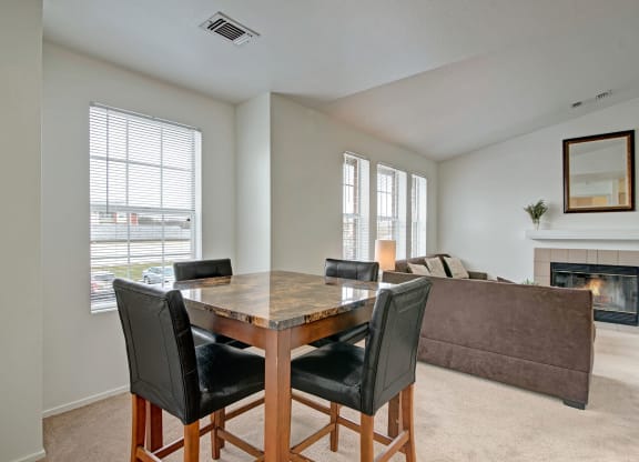 A modern living area with beige walls and carpet features a wooden dining table with four black chairs near a window. A cozy sofa faces a lit fireplace at St. James Place Apartments.