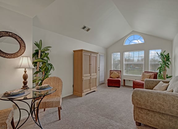 Cozy living room with vaulted ceiling, featuring a beige sofa, two red armchairs, a wooden cabinet, and large windows letting in natural light at St. James Place Apartments.
