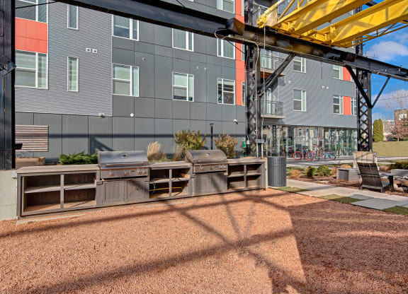 Outdoor area with two metal grills under a modern structure, adjacent to a gray building. Bicycles and lounge chairs are visible. Bright, sunny day at Stitchweld Apartments.