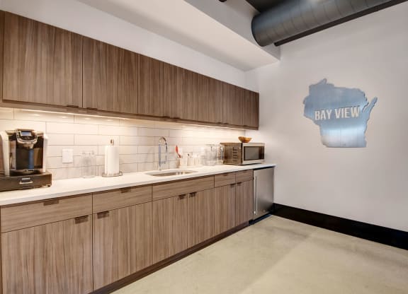 Modern kitchen with wooden cabinets, white countertops, and a stainless steel sink. A sign shaped like Wisconsin says "eat well" on the wall, creating a welcoming vibe at Stitchweld Apartments.