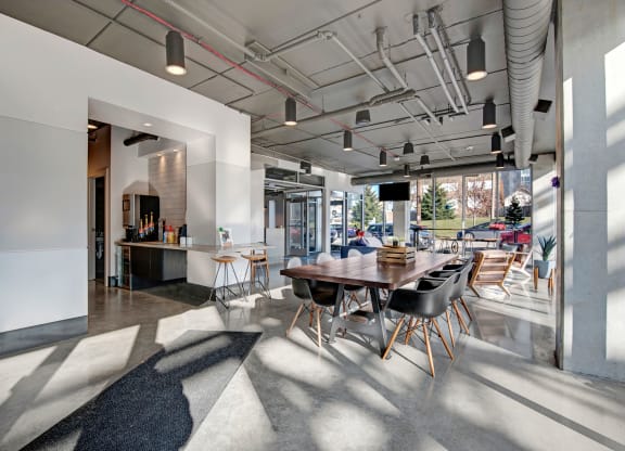 Modern cafe interior with large windows and natural light. Features a long wooden table with black chairs, a coffee station, and minimalist decor at Stitchweld Apartments.