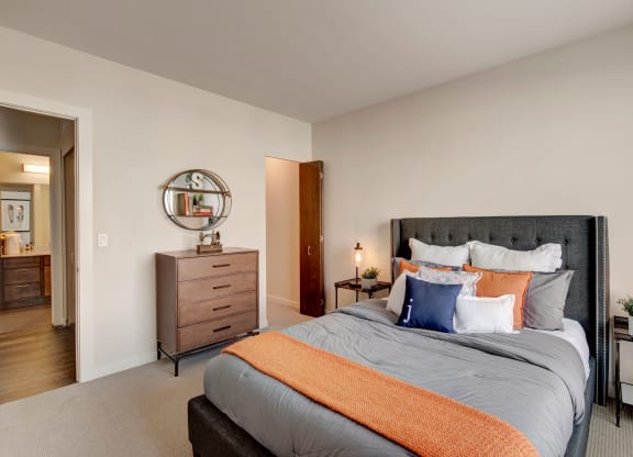 Modern bedroom with a plush gray bed, layered pillows, and an orange throw blanket. A wooden dresser and round mirror are on the left at Stitchweld Apartments.