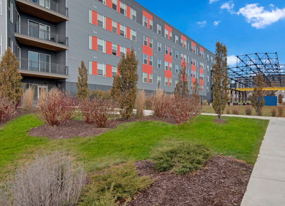 Modern apartment building with gray and red panels, surrounded by landscaped greenery. A pathway and metal structure are visible under a clear blue sky at Stitchweld Apartments.