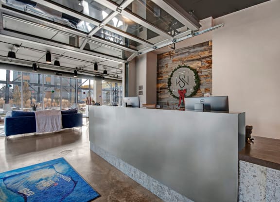 Modern reception desk with minimalist design, featuring two computers. A logo with a holiday wreath is on the wall. Large windows and blue couches add a welcoming ambiance at Stitchweld Apartments.