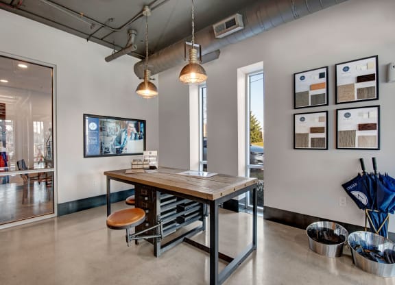 Modern office lobby with industrial decor. A wooden table and stool set is centered under two pendant lights. Sample boards and umbrellas line the walls, creating a welcoming atmosphere at Stitchweld Apartments.