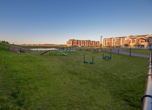 A grassy dog park with agility equipment, fenced and bordered by The Park Apartments, a row of red-brick apartments in the background.