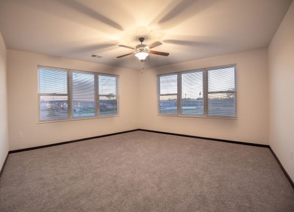 Spacious empty room at the Park Apartments with beige walls and carpet, ceiling fan, and large windows with blinds showing a sunset sky, creating a serene atmosphere.