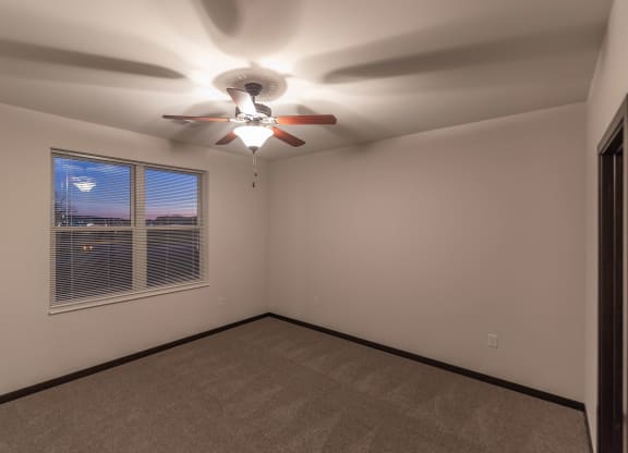 Empty beige room with a beige carpet. A ceiling fan with wooden blades is lit overhead. A window with blinds shows a twilight sky at The Park Apartments.