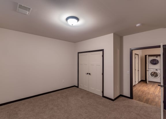 An empty room with beige walls and carpet flooring features a closed double door and an open door leading to a laundry area with a stacked washer and dryer at The Park Apartments.