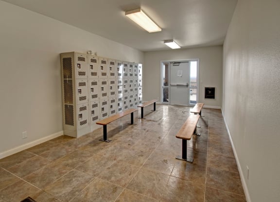 A clean, empty mail room here at Confluence at the Harvest Hills Apartment Homes with beige floor tiles, metal lockers lining the left wall, and wooden benches in the middle. Overhead fluorescent lighting.