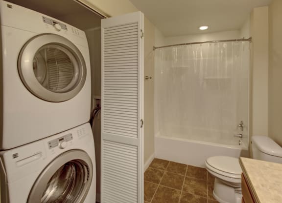 Compact laundry and bathroom space here at Confluence at the Harvest Hills Apartment Homes with stacked washer and dryer on the left, white bathtub with a curtain, toilet, and tiled floor. Clean and functional.