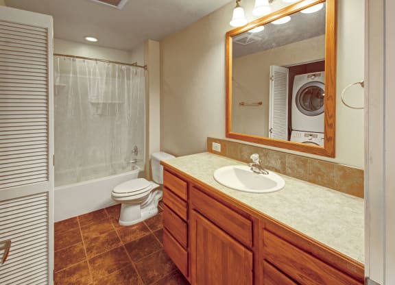 Compact bathroom here at Confluence at the Harvest Hills Apartment Homes with wooden cabinets below a sink and mirror. White toilet and shower with curtain on the left, stacked washer and dryer visible. Warm lighting.
