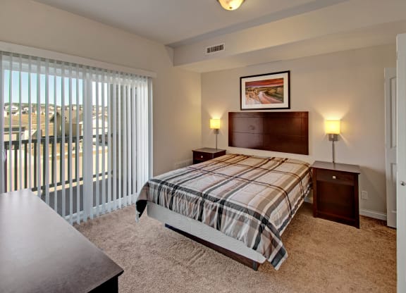 A cozy bedroom here at Confluence at the Harvest Hills Apartment Homes with a plaid-patterned bed, flanked by two nightstands and wall lamps. Large window with vertical blinds offers natural light.