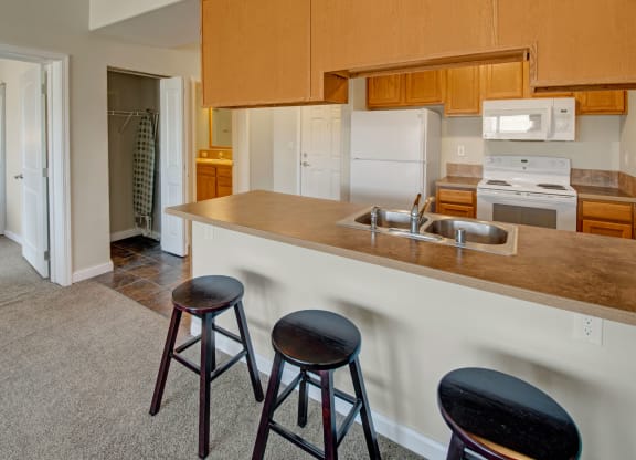 A cozy kitchen here at Confluence at the Harvest Hills Apartment Homes with wooden cabinets, a white stove, fridge, and microwave. A bar counter with three stools faces the sink, conveying a welcoming vibe.