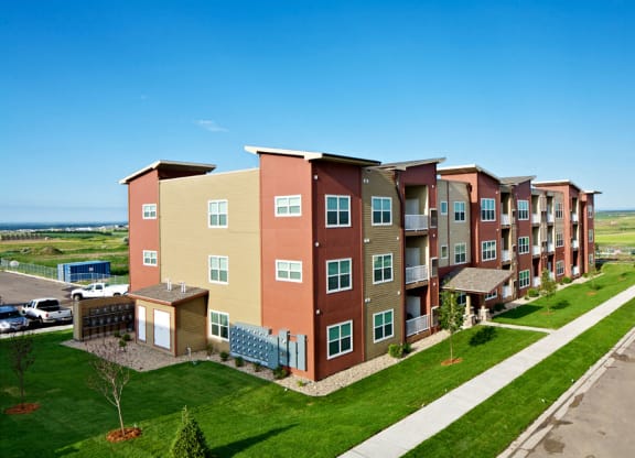 Modern three-story Confluence at the Harvest Hills Apartment Homes building with red and beige siding, bordered by a green lawn and sidewalk, set against a rural landscape under a clear blue sky.