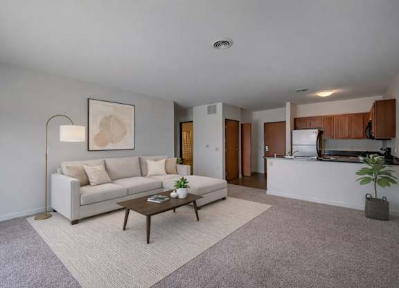 A bright living room here at Latitude featuring a cozy beige sectional sofa, light neutral rug, wooden coffee table, and floor lamp, creating an open, inviting space connected to a warm kitchen with wood cabinetry.
