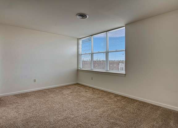 A bright bedroom here at Latitude featuring large sunlit windows, soft carpet flooring, and a spacious empty layout, creating a clean, open, and inviting space ready for personalization.