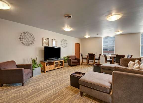 A welcoming resident lounge here at Latitude featuring warm brown seating, a large TV console, and dining tables by the windows, creating a comfortable and inviting space for relaxing or gathering.