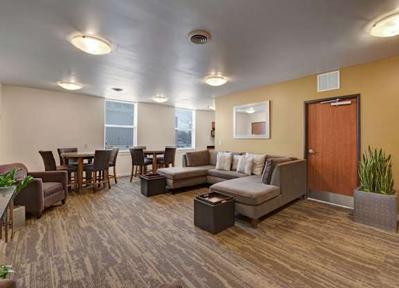 A comfortable resident lounge here at Latitude featuring a cozy sectional sofa, warm accent wall, and dining tables by the windows, creating a relaxing and inviting space for gathering or unwinding.