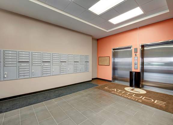 A welcoming mailroom lobby here at Latitude featuring a warm orange accent wall, secure gray mailboxes, and modern elevators, creating a clean and convenient space for residents.