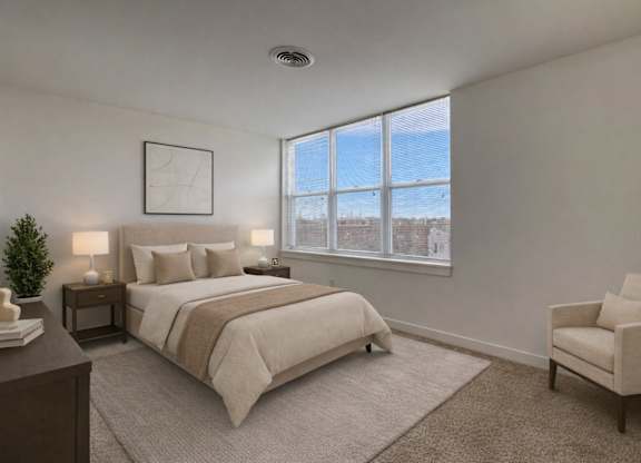 A bright bedroom here at Latitude featuring neutral bedding, dark wood nightstands, and a soft area rug, complemented by a cozy accent chair and large sunlit windows creating a calm, welcoming atmosphere.