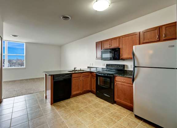 A bright kitchen here at Latitude featuring warm wood cabinetry, black appliances, and tiled flooring, opening to a spacious, inviting living area with soft carpet and a large sunlit window.