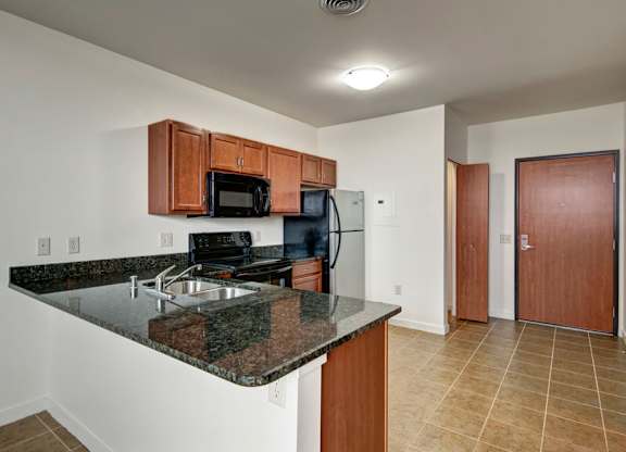 A bright kitchen here at Latitude featuring rich wood cabinetry, black appliances, and a spacious granite breakfast bar, complemented by warm tile flooring and a clean, open, welcoming layout.