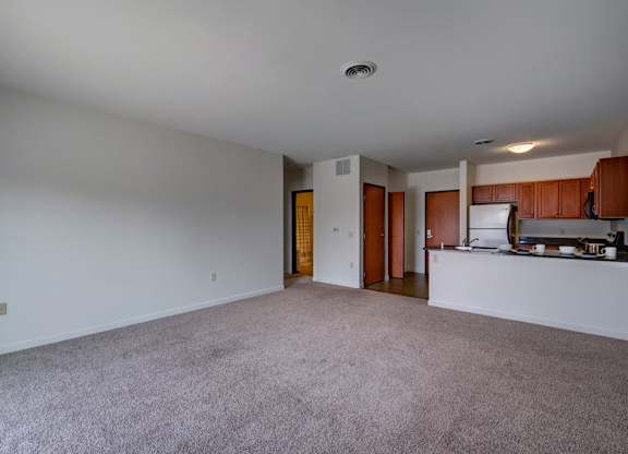 A spacious living room here at Latitude featuring soft neutral carpeting, an open layout flowing into the kitchen with warm wood cabinetry, and bright walls that create a clean, airy, welcoming atmosphere.
