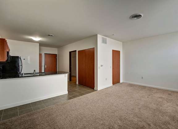A spacious living room here at Latitude featuring soft neutral carpeting, bright walls, and an open layout leading into the kitchen with warm wood cabinetry, creating a clean, comfortable, and welcoming atmosphere.
