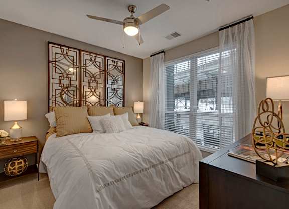 A cozy bedroom here at River House featuring a white bed with warm gold pillows, dark wood nightstands, and sheer curtains over a large window, creating a bright, inviting, and relaxing space.
