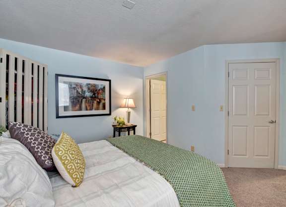 A bright bedroom here at River’s Cove featuring a plush bed with green bedding, soft carpet, and calming blue walls. A side table with lamp and framed artwork adds a warm, welcoming touch.