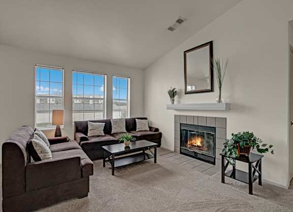 A cozy living room here at St. James Place with soft beige carpet, cream walls, and a warm corner fireplace. A comfortable seating area sits beside tall windows that bring in natural light, creating a bright and inviting atmosphere.