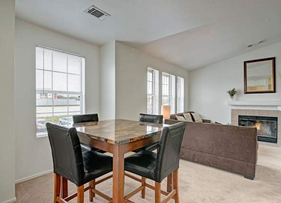 A bright dining area here at St. James Place with soft beige carpet, large windows, and a modern dining table. A nearby fireplace and open living space create a warm and inviting atmosphere.