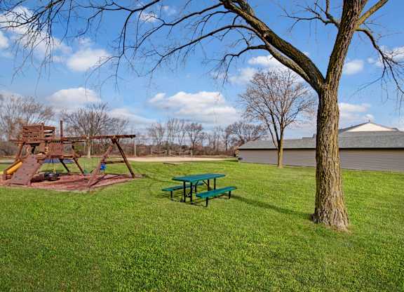 A spacious outdoor playground here at St. James Place with a wooden play structure, swings, and a grassy open area. A nearby picnic table and mature trees create a relaxing and family-friendly atmosphere.