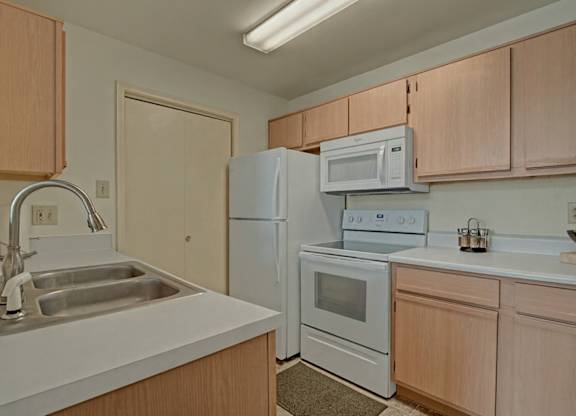 A bright kitchen here at St. James Place with light wood cabinets, white countertops, and white appliances. A double-basin sink and overhead lighting create a clean and functional space with a warm, inviting feel.