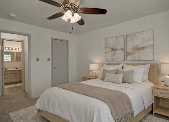 A spacious bedroom here at St. James Place with soft beige carpet, cream walls, and a ceiling fan for added comfort. A connected bathroom and warm lighting create a convenient and inviting atmosphere.