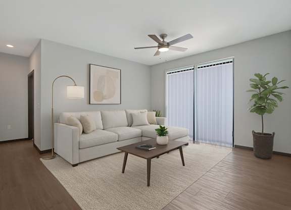 A bright living room here at The Lafayette featuring a cream sectional sofa, warm wood coffee table, light rug, sliding glass doors with vertical blinds, ceiling fan, and a tall green plant in the corner.