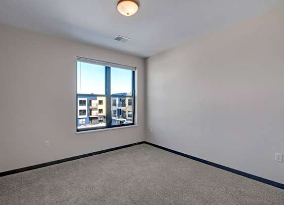 A bright bedroom here at The Lafayette with soft beige carpet, light walls, and dark trim. A large window brings in natural light and offers a pleasant view of the surrounding community buildings.