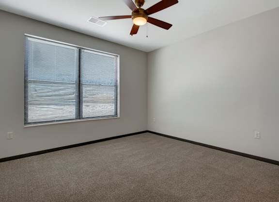A comfortable bedroom here at The Lafayette with soft beige carpet, light walls, and dark trim. A large window with blinds brings in natural light, and a ceiling fan adds comfort and airflow.