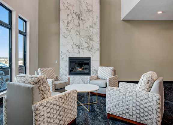A cozy lounge here at The Lafayette with patterned accent chairs, a round table, and a tall marble fireplace. A large window brings in natural light, creating a warm, inviting space to relax.