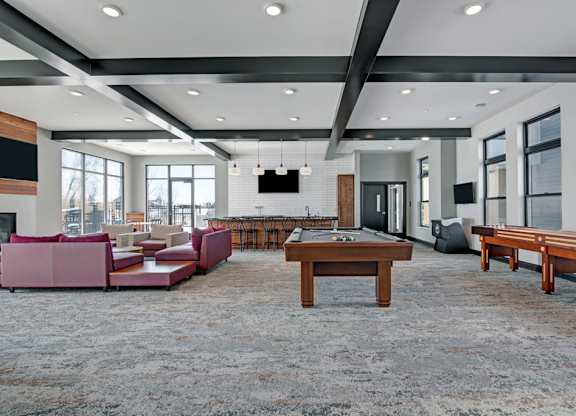 A spacious community lounge here at The Lafayette with a pool table, shuffleboard, and comfortable seating. A large kitchen island, bar seating, and bright windows create a lively, welcoming space for entertaining.